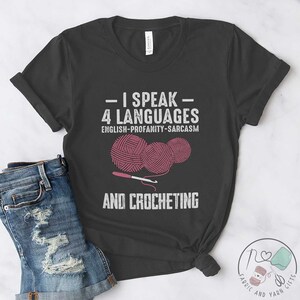 May include: A dark gray t-shirt with the text "I speak 4 languages: English-Profanity-Sarcasm and crocheting" and a graphic of three pink yarn balls and a crochet hook.