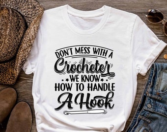 May include: White t-shirt with black text that reads "Don't mess with a crocheter we know how to handle a hook". The text is in a stylized font. A crochet hook is pictured below the text.
