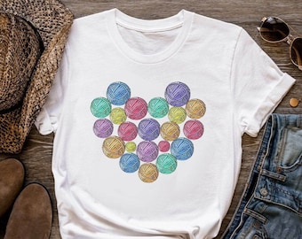 Puede incluir: Camiseta blanca con un diseño de corazón de lana de colores. El corazón está formado por muchas pequeñas bolas de lana de diferentes colores.