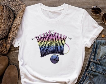 Puede incluir: Camiseta blanca con un gráfico de un proyecto de tejido de color arcoíris con agujas y una bola de hilo.