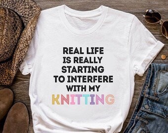 Puede incluir: Camiseta blanca con el texto "Real life is really starting to interfere with my knitting" en negro. La palabra "knitting" está en colores arcoíris.