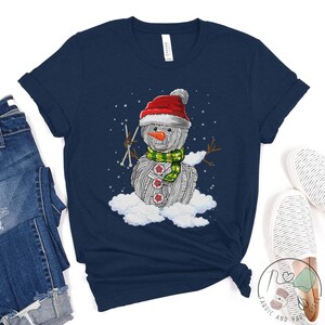 Pu&ograve; includere: Una maglietta blu navy con un pupazzo di neve di lana bianca che indossa un cappello di Babbo Natale rosso e una sciarpa verde. Il pupazzo di neve tiene in mano i ferri da maglia e si trova su una nuvola.