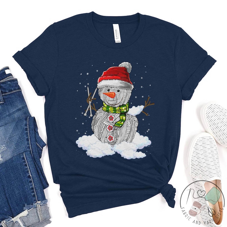Pu&ograve; includere: Una maglietta blu navy con un pupazzo di neve di lana bianca che indossa un cappello di Babbo Natale rosso e una sciarpa verde. Il pupazzo di neve tiene in mano i ferri da maglia e si trova su una nuvola.