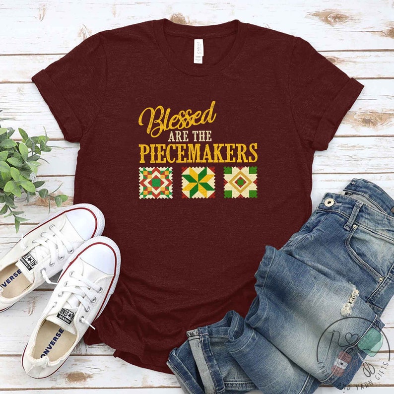 May include: A maroon t-shirt with the text "Blessed Are The Peacemakers" in gold lettering. Three colorful quilt squares are below the text.
