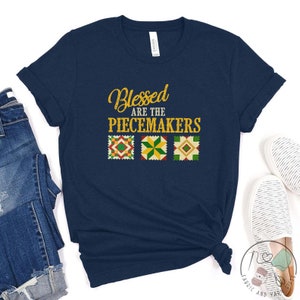 May include: Navy blue t-shirt with the text "Blessed are the Peacemakers" in gold lettering. Three colorful quilt squares are below the text.