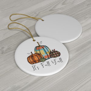 Può includere: Ciondolo in ceramica con sfondo bianco e un disegno di tre zucche. Le zucche sono decorate con motivi a quadri in tonalità di arancione, blu, verde e rosso. Il testo "It's Fall Y'all" è scritto in nero sotto le zucche.