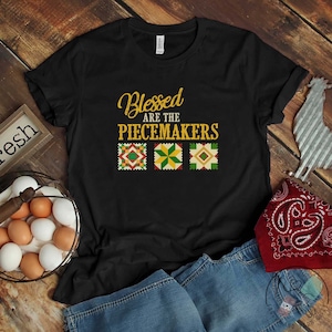 May include: Black t-shirt with the text "Blessed are the Peacemakers" in gold lettering. Three colorful quilt squares are below the text.