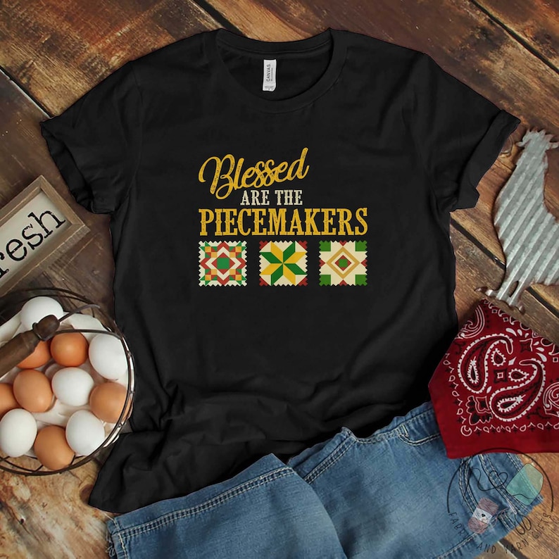 May include: Black t-shirt with the text "Blessed are the Peacemakers" in gold lettering. Three colorful quilt squares are below the text.