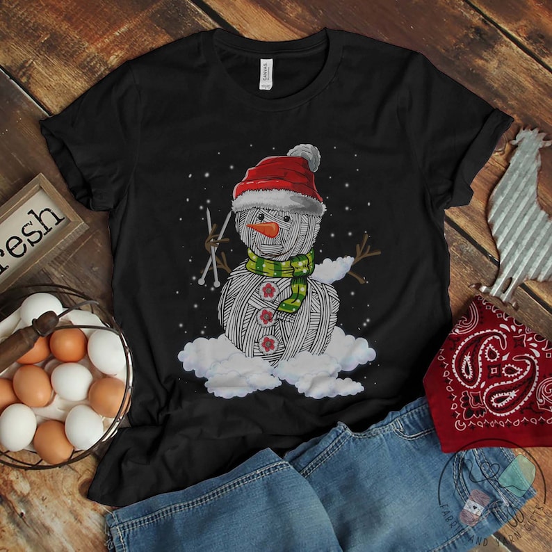 Pu&ograve; includere: Maglietta nera con un pupazzo di neve di lana bianca che indossa un cappello di Babbo Natale rosso e una sciarpa verde. Il pupazzo di neve tiene in mano gli aghi da maglia.