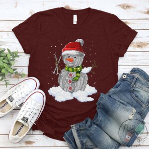 Pu&ograve; includere: Una maglietta color granata con un pupazzo di neve di lana bianca che indossa un cappello di Babbo Natale rosso e una sciarpa verde. Il pupazzo di neve tiene in mano due ferri da maglia ed &egrave; seduto su una nuvola.