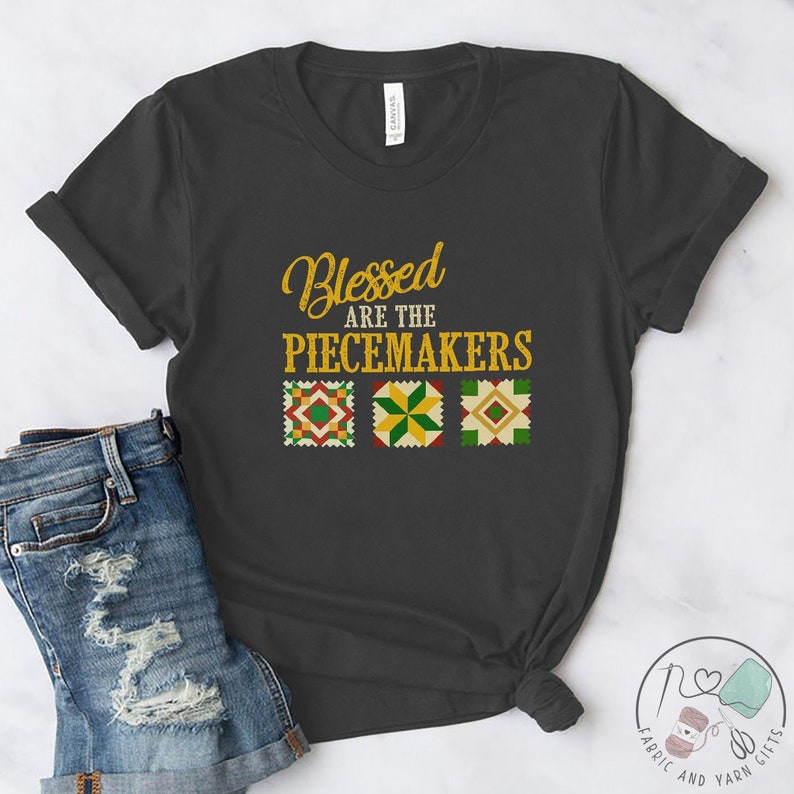 May include: A dark gray t-shirt with the text "Blessed are the Peacemakers" in gold lettering. Three colorful quilt squares are below the text.