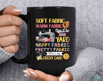 May include: Black coffee mug with a white design featuring a sewing machine, fabric scraps, and the text "Soft Fabric, Warm Fabric, Buy it by the Yard, Happy Fabric, Pretty Fabric, Here's my Credit Card".