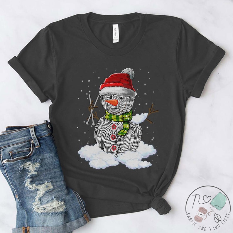 Pu&ograve; includere: Una maglietta grigia con un pupazzo di neve fatto di lana che indossa un cappello da Babbo Natale rosso e una sciarpa verde. Il pupazzo di neve tiene in mano gli aghi da maglia ed &egrave; circondato da nuvole bianche.