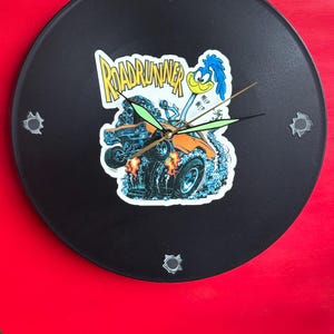 May include: A black Road Runner clock with a cartoon image of the Road Runner and a car. The clock hands are green and the hour markers are silver. The clock is mounted on a red surface.