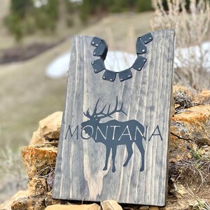 以下が含まれることがあります： 黒いシルエットのヘラジカと黒い文字で「MONTANA」と書かれた灰色の木製看板。看板の上部には装飾的な金属製の縁取りがある。
