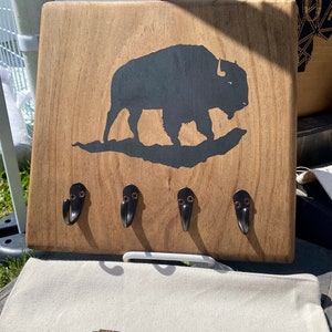 May include: Wooden key holder with a black silhouette of a buffalo and four metal hooks. The key holder has a natural wood finish.