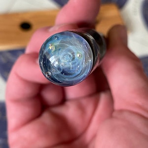 May include: A close-up of a glass pen with a swirling blue and white design. The pen has a clear glass body and a silver metal cap. There are small silver flecks embedded in the glass.