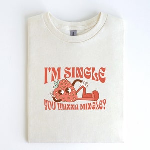 May include: A white t-shirt with a red strawberry graphic and the text "I'm single, you wanna mingle?"
