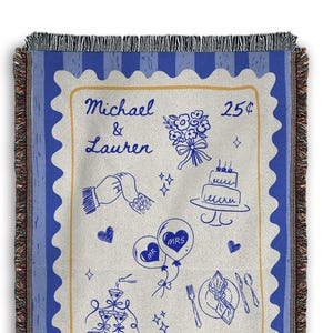 May include: A woven throw blanket with a blue and cream design. The centre features a stamp-like illustration with wedding-themed drawings and the names "Michael & Lauren." The border is blue with a fringed edge.
