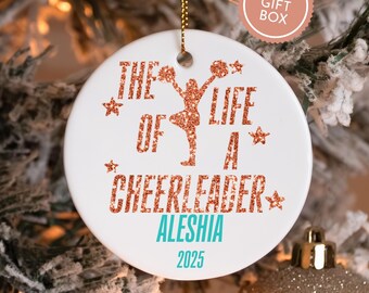 May include: White ceramic ornament with the text "THE LIFE OF A CHEERLEADER" in glittery orange, with a cheerleader graphic. The name "ALESHIA" and the year "2025" are in teal. Includes a gold string for hanging.