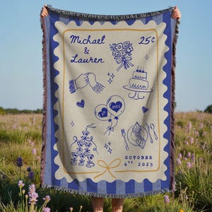 May include: A beige and blue woven blanket with a wedding design. The blanket features illustrations of a bouquet, cake, champagne glasses, and balloons. The names "Michael & Lauren" and the date "October 6th 2025" are also displayed.