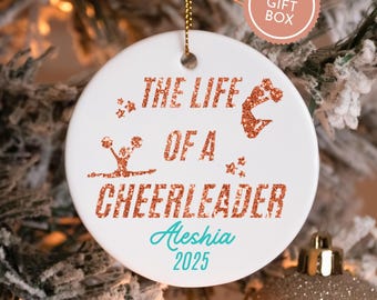 May include: White ceramic Christmas ornament with gold string. The ornament reads "THE LIFE OF A CHEERLEADER" in glittery copper lettering, with the name "Aleshia" and the year "2025" in teal. A "FREE GIFT BOX" sticker is in the background.