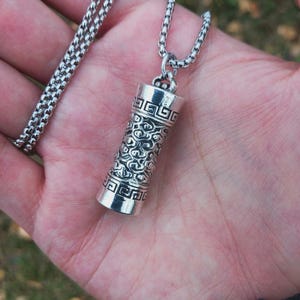May include: A silver-colored pendant necklace. The pendant is cylindrical with intricate engraved patterns. The chain is a box chain style. The necklace is displayed on a hand.