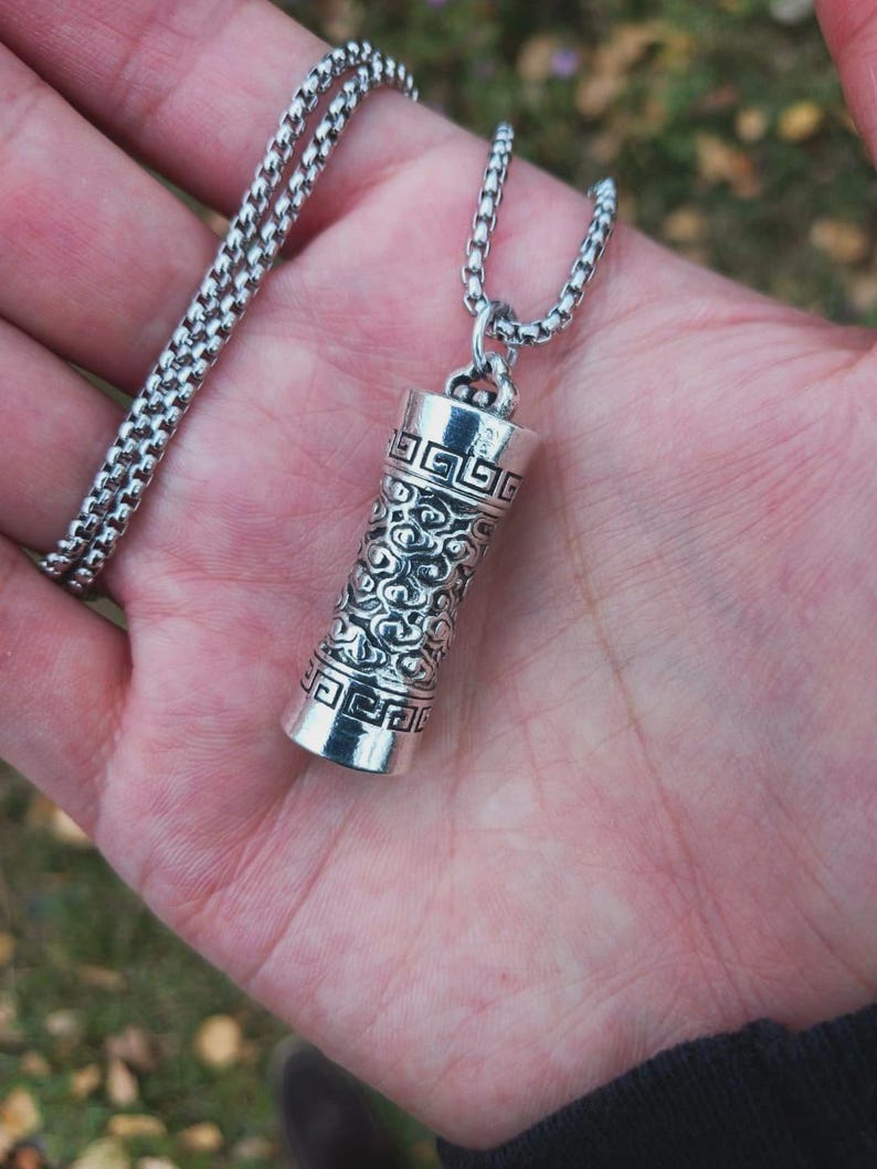 May include: A silver-colored pendant necklace. The pendant is cylindrical with intricate engraved patterns. The chain is a box chain style. The necklace is displayed on a hand.