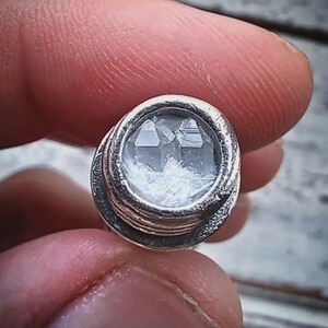 May include: A small, round, silver-colored object with a clear, faceted crystal center. The crystal appears to have a cluster of smaller crystals inside. The object is held between two fingers, suggesting it is a small decorative item or jewelry component.
