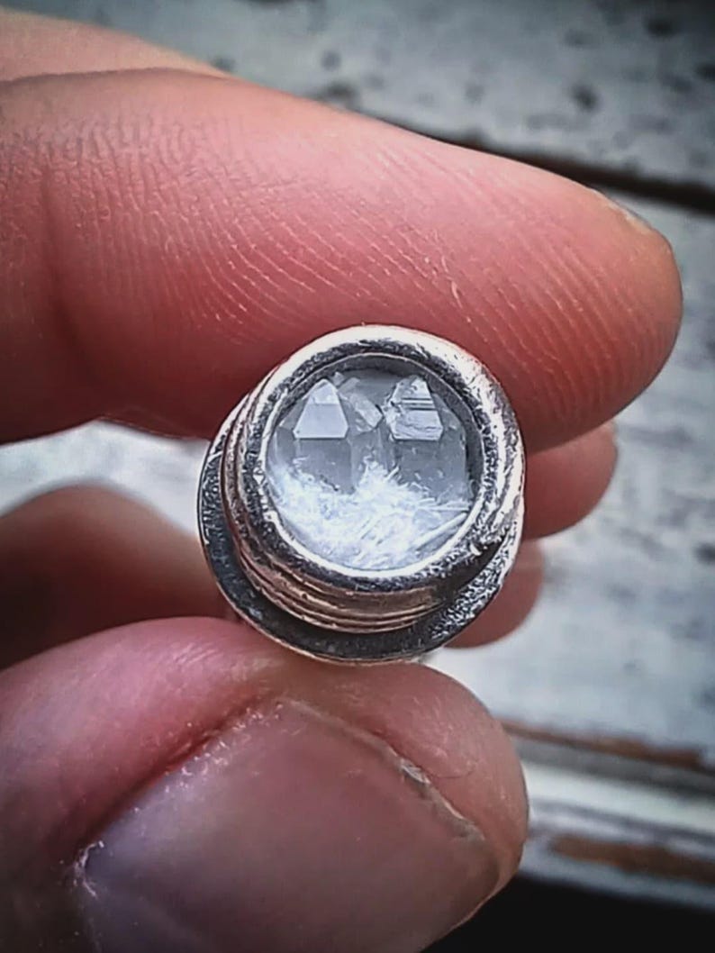May include: A small, round, silver-colored object with a clear, faceted crystal center. The crystal appears to have a cluster of smaller crystals inside. The object is held between two fingers, suggesting it is a small decorative item or jewelry component.