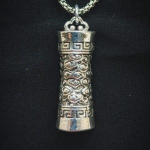 May include: Silver-toned cylindrical pendant with intricate cloud and geometric patterns. The pendant hangs from a silver chain. The design features detailed textures and a polished finish, creating a striking visual appeal. The background is a solid black.