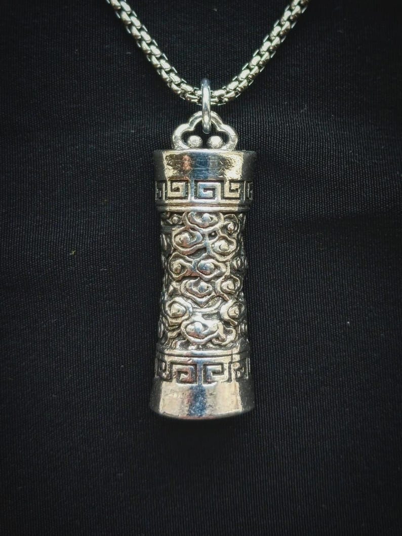 May include: Silver-toned cylindrical pendant with intricate cloud and geometric patterns. The pendant hangs from a silver chain. The design features detailed textures and a polished finish, creating a striking visual appeal. The background is a solid black.