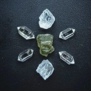 May include: An overhead shot of several crystals arranged on a dark surface. Six clear quartz crystals are arranged in a circle around a central, olive-green crystal. The quartz crystals are faceted and have a crystalline structure.