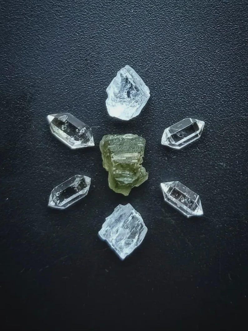 May include: An overhead shot of several crystals arranged on a dark surface. Six clear quartz crystals are arranged in a circle around a central, olive-green crystal. The quartz crystals are faceted and have a crystalline structure.