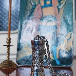 May include: A silver-colored pendant necklace with a cylindrical design featuring intricate patterns. The pendant hangs from a chain. The background includes a religious icon and a brass candlestick.