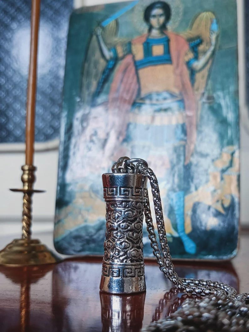 May include: A silver-colored pendant necklace with a cylindrical design featuring intricate patterns. The pendant hangs from a chain. The background includes a religious icon and a brass candlestick.