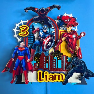 May include: A cake topper featuring a cityscape with a red glitter finish and the name Liam.  The topper also includes five superhero figures: Spiderman, Iron Man, Captain America, Superman, and Black Panther. The number 3 is displayed in red and yellow.
