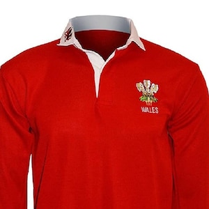 May include: A red long-sleeved rugby jersey with a white collar and a white embroidered Welsh dragon crest with the word "WALES" underneath.