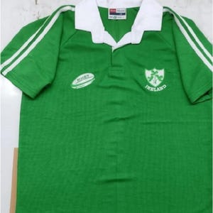 May include: Green rugby jersey with white stripes on the sleeves. The jersey has a white collar and a white embroidered rugby ball on the left side. The right side of the jersey has an embroidered crest with the words "Ireland" underneath.
