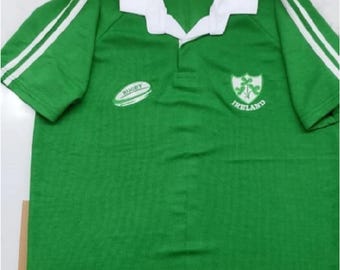 May include: Green rugby jersey with white stripes on the sleeves. The jersey has a white collar and a white embroidered rugby ball on the left side. The right side of the jersey has an embroidered crest with the words "Ireland" underneath.
