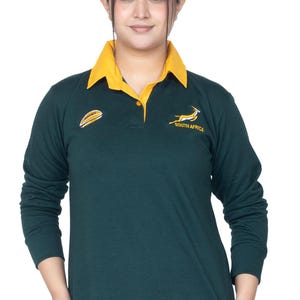 以下が含まれることがあります： 胸に南アフリカの春bokのロゴが付いた、濃い緑色の長袖のラグビーシャツ。ロゴは白い春bokと「SOUTH AFRICA」の文字で構成されています。