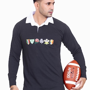 Könnte beinhalten: Ein dunkelgraues Rugby-Langarmshirt mit weißem Kragen und gestickten Emblemen. Das Shirt wird mit einer blauen Jeans kombiniert. Ein brauner und weißer Football wird in den Händen gehalten.
