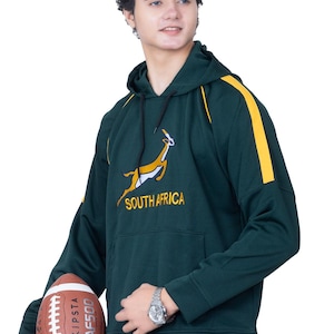 May include: A green hooded sweatshirt with yellow accents and a South Africa rugby team logo featuring a springbok. The sweatshirt has a kangaroo pocket and a drawstring hood. A brown and white football is held in the left hand of the person wearing the sweatshirt.