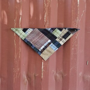 May include: A patchwork bandana in a triangular shape, featuring a variety of plaid and patterned fabrics in shades of blue, green, brown, and cream. The bandana is displayed against a reddish-brown background.