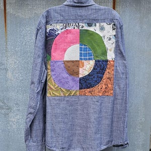 May include: A blue button-down shirt with a patchwork design on the back. The patchwork is a circle divided into colorful sections, including pink, green, blue, and orange patterns. The shirt is hanging on a metal bar.