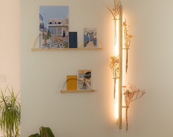 May include: A wooden wall mounted light fixture with a vertical design. The fixture has three glass tubes that hold dried flowers and plants. The light fixture is illuminated with warm white LED lights. The fixture is mounted on a white wall with two wooden shelves holding framed artwork.