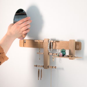 Könnte beinhalten: Ein hölzerner Schmuckorganizer mit einem Spiegel, einem Regal und mehreren Stangen zum Aufhängen von Halsketten, Armbändern und Ohrringen. Der Organizer ist an einer weißen Wand montiert.