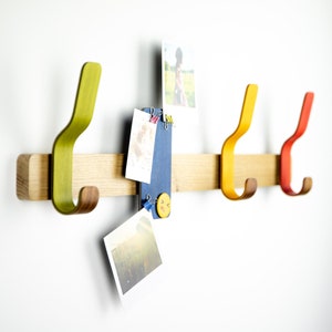 May include: A wooden wall-mounted coat rack with four hooks. The hooks are different colors: green, yellow, orange, and red. There are photos and a small blue board with a smiley face button pinned to the rack.
