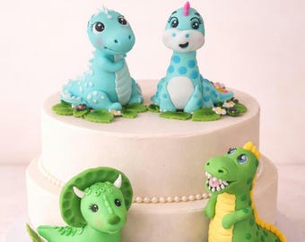 May include: A two-tiered white cake decorated with four cartoon dinosaur figurines. Two blue dinosaurs with white spots and two green dinosaurs with yellow accents sit on the cake, surrounded by green leaves and small white flowers.