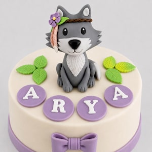 Könnte beinhalten: Ein dekorierter Kuchen mit einer grauen Wolfsfigur mit Blumenkranz. Der weiße Kuchen hat lila Akzente, darunter eine Schleife und den Namen "ARYA" in lila Kreisen. Grüne Blattdekorationen vervollständigen das Design.